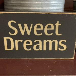 May include: A black wooden sign with distressed edges and a white background. The sign reads "Sweet Dreams" in gold lettering.
