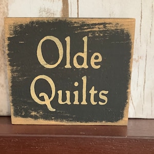 May include: A black wooden sign with white lettering that reads "Olde Quilts".