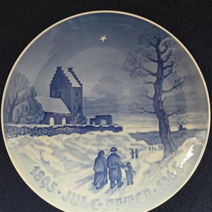 Może przedstawiać: Błękitno-biały talerz ceramiczny z zimową sceną przedstawiającą kościół, rodzinę idącą i gwiazdę na niebie. Na brzegu talerza widnieje napis "1895. JUL. C. AFTEN. 1965".