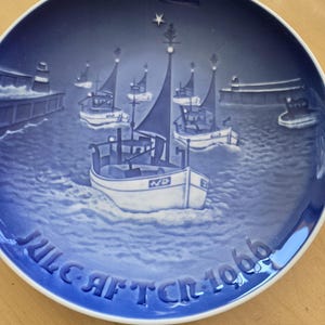 May include: A blue and white Christmas plate with a scene of fishing boats sailing into a harbor at night. The plate is decorated with a star and a Christmas tree. The text "JUL. AFT. CR. 1966" is printed on the bottom of the plate.