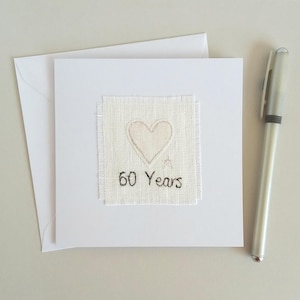 May include: A white greeting card with a stitched heart design and the text "60 Years". The card is accompanied by a white envelope and a silver pen. The heart is embroidered on a square of textured fabric.