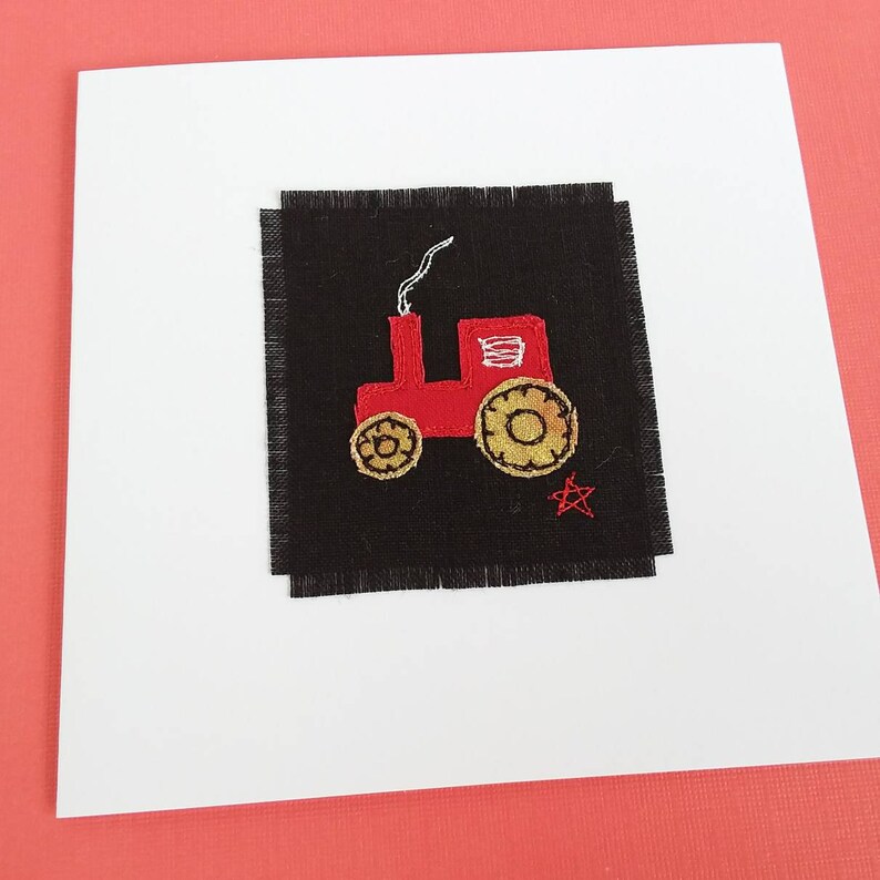 Puede incluir: Una tarjeta de felicitaci&oacute;n hecha a mano con un tractor rojo bordado sobre un fondo de tela negra. El tractor tiene una columna de humo blanca, una ventana amarilla y ruedas doradas. Una estrella roja est&aacute; bordada debajo del tractor.