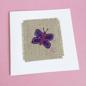May include: A white card with a square piece of beige linen fabric in the center. The fabric has a purple butterfly with a pink body and black antennae stitched onto it.