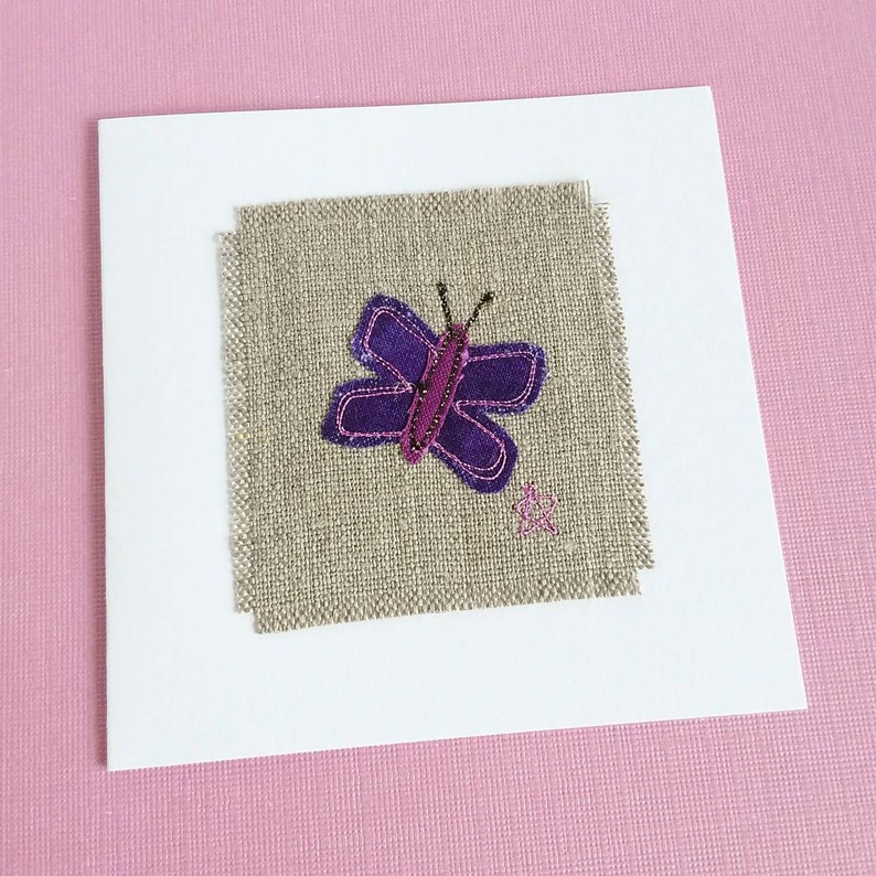 May include: A white card with a square piece of beige linen fabric in the center. The fabric has a purple butterfly with a pink body and black antennae stitched onto it.