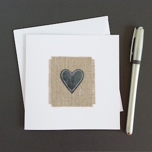 May include: A white card with a burlap square with a black heart in the center. The card is on a black surface with a silver pen.