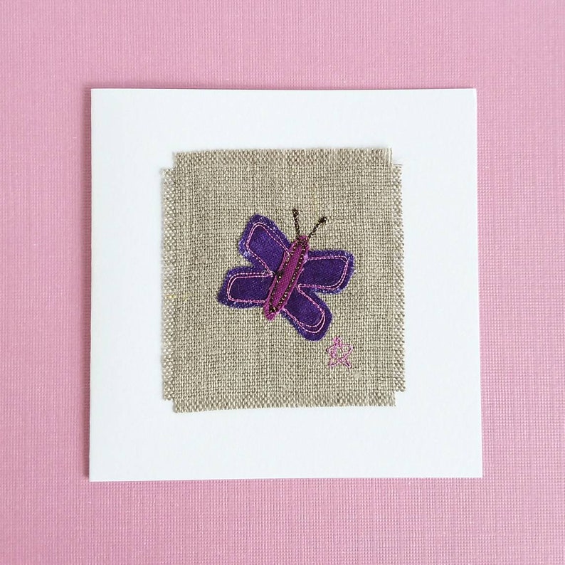 May include: A white card with a purple butterfly embroidered on a piece of beige linen fabric.