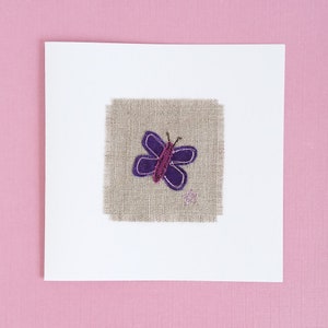 May include: A white card with a purple butterfly embroidered on a piece of beige fabric.