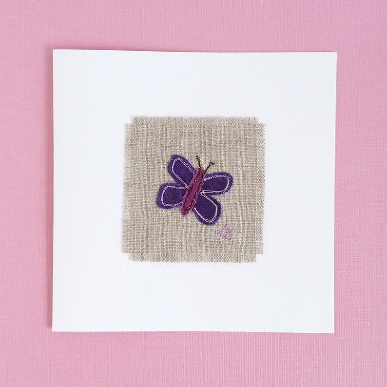 May include: A white card with a purple butterfly embroidered on a piece of beige fabric.