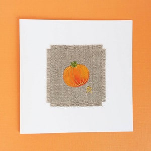 May include: A handmade card with a small embroidered pumpkin on a beige linen background. The pumpkin is orange with a green stem and a gold star.