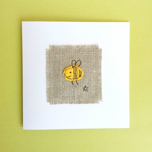 May include: A white card with a small piece of hessian fabric sewn onto it. The hessian fabric has a yellow embroidered bee with black details.