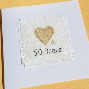 May include: A white greeting card with a square, textured patch featuring a gold heart with a swirl design, a gold star, and the text "50 Years" embroidered in black thread. The card is on a yellow background.