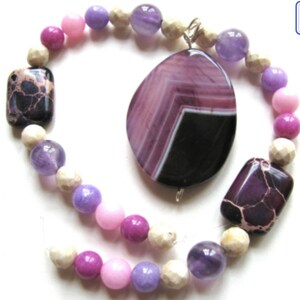 May include: A purple and black agate gemstone pendant with a silver chain, surrounded by a beaded bracelet with white, pink, and purple beads.