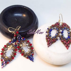 Può includere: Un paio di orecchini fatti a mano a forma di goccia. Gli orecchini presentano perline rosse, blu, oro e argento, con ganci dorati. Esposti su una superficie bianca, accanto a una ciotola nera.