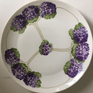 May include: White ceramic plate with a floral design. The plate features clusters of purple flowers with green leaves, connected by thin gold lines. The flowers are arranged around the plate's edge and center.