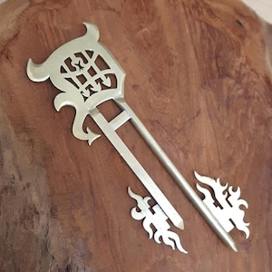 Op de afbeelding: Twee messing haaraccessoires. Eén is een lange haarspeld met een decoratieve bovenkant en een kleiner bijpassend accentstuk. De stukken hebben een sierlijk, vintage ontwerp met een vlamachtig motief.