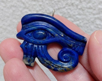 May include: A blue Eye of Horus pendant carved from stone. The pendant features intricate details, including an eye, eyebrow, and stylized markings. A small silver loop is attached for hanging. The stone has a polished finish.