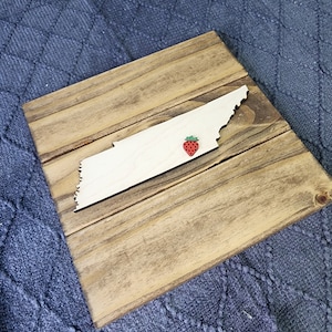 May include: A wooden cutout of the state of Tennessee with a red and green strawberry decoration on the state.