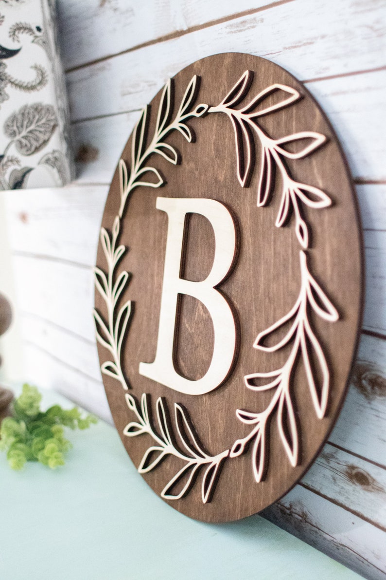May include: A round wooden sign with a dark brown finish. The sign features a large, off-white letter "B" in the center, surrounded by a laser-cut leaf design. The sign is approximately 12 inches in diameter.