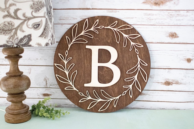 May include: A round wooden sign with a dark brown finish. The sign features a large, light-colored letter "B" in the center, surrounded by a decorative wreath of leaves. The leaves are cut out of the wood, creating a contrasting design.