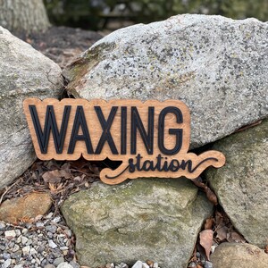 May include: A wooden sign with the words "Waxing Station" in black lettering. The sign is sitting on a pile of rocks.