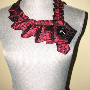 May include: A red and black patterned tie necklace with a black button in the center. The necklace is on a gray mannequin.