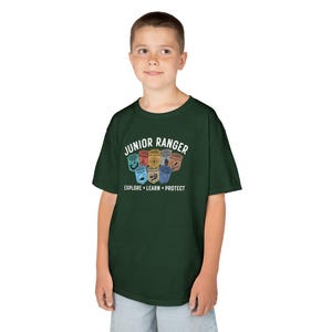 May include: A dark green t-shirt with a white graphic that says "Junior Ranger" and features a collection of colorful badges. The text below the badges says "Explore. Learn. Protect."