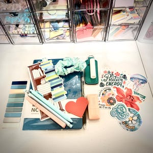 May include: A collection of stationery and accessories, including pens, stickers, a scrunchie, and a notebook. The items are arranged on a white surface, with a clear organizer in the background. Stickers include the phrases "Positive Energy" and "Stay Humble".