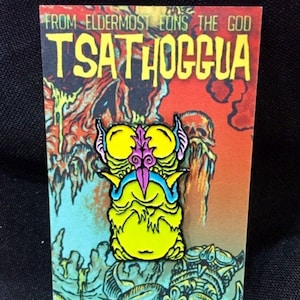 May include: A yellow enamel pin with a cartoon monster with pink and purple details. The pin is on a card with the text "From Eldermost Eons The God Tsathhogua" and "A Nick the Hat product".