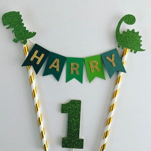 May include: Green and gold glitter cake topper with a dinosaur, a banner that says "HARRY", and the number "1".