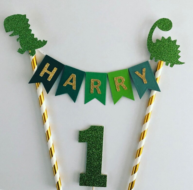 May include: Green and gold glitter cake topper with a dinosaur, a banner that says "HARRY", and the number "1".