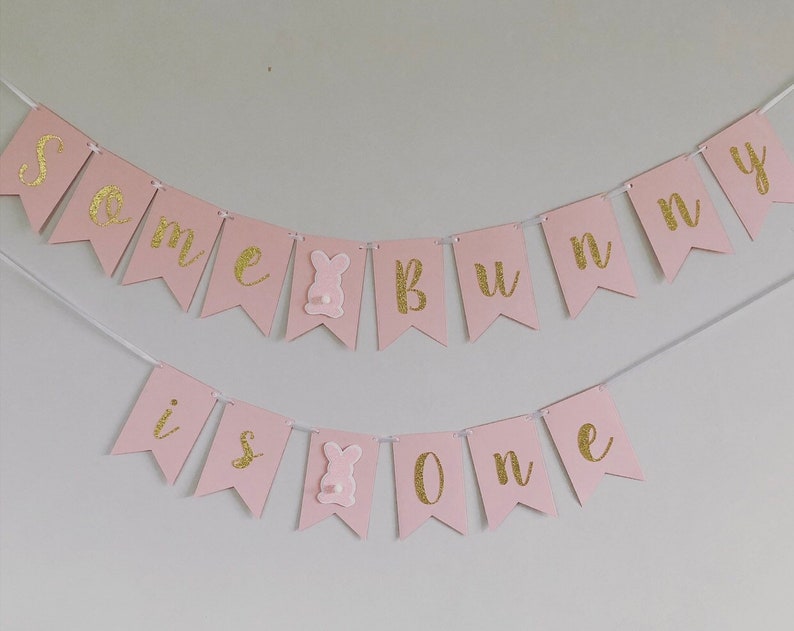 Op de afbeelding: Roze en gouden banner met de tekst "Some bunny is one". De banner is gemaakt van vilt en heeft aan elke kant een konijnenuitsparing.
