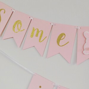 Op de afbeelding: Roze en gouden banner met het woord "een" in gouden glitterletters. De banner is versierd met een roze konijn.