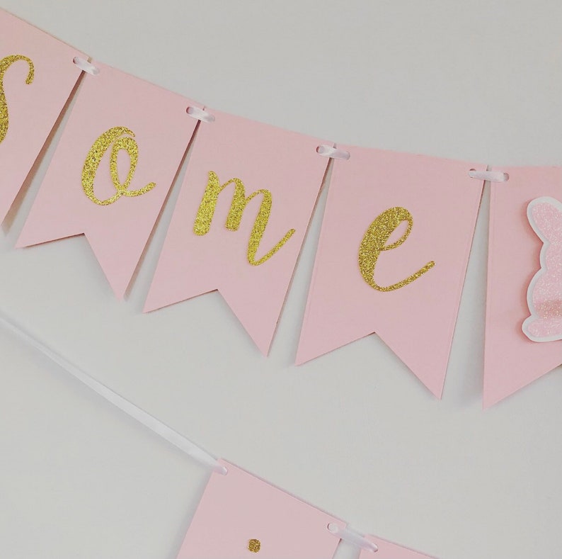 Op de afbeelding: Roze en gouden banner met het woord "een" in gouden glitterletters. De banner is versierd met een roze konijn.