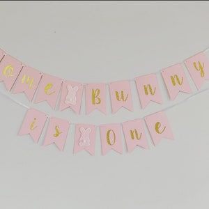 Op de afbeelding: Roze en gouden banner met de tekst "Some bunny is one". De banner is gemaakt van papier en heeft een touwtje eraan bevestigd. Er zitten twee konijnenuitsnijdingen op de banner.