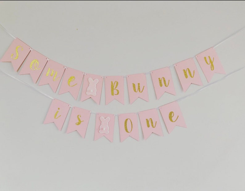 Op de afbeelding: Roze en gouden banner met de tekst "Some bunny is one". De banner is gemaakt van papier en heeft een touwtje eraan bevestigd. Er zitten twee konijnenuitsnijdingen op de banner.