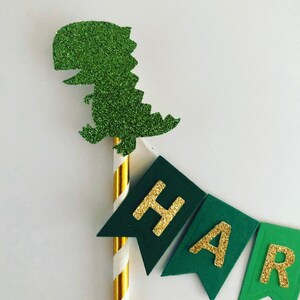 May include: Green glitter dinosaur cake topper with a gold glitter banner that says "Happy".