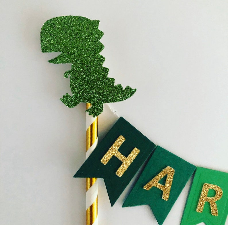 May include: Green glitter dinosaur cake topper with a gold glitter banner that says "Happy".