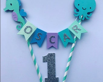 Puede incluir: Un conjunto de adornos para tartas con un caballito de mar, un pulpo y una pancarta que dice "OSCAR" con un número 1 de purpurina plateada. La pancarta está hecha de papel azul, morado y verde.