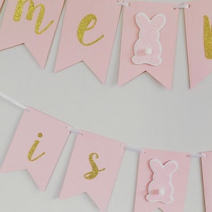 Op de afbeelding: Roze en gouden banner met de woorden "Home is" en een konijn aan elk uiteinde.