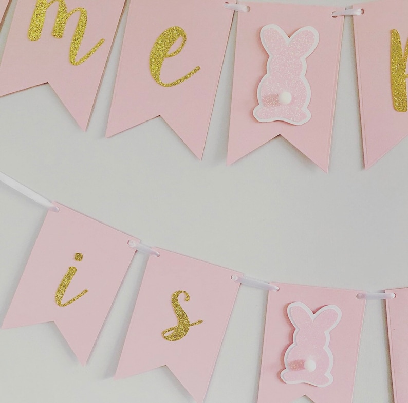 Op de afbeelding: Roze en gouden banner met de woorden "Home is" en een konijn aan elk uiteinde.