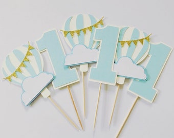 May include: Set of six blue and white hot air balloon cake toppers with gold glitter flags and clouds. The toppers are on wooden sticks and spell out the number "one".
