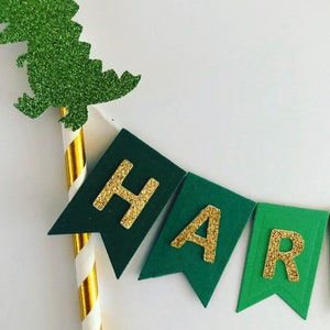May include: Green and gold glitter banner with the word "Happy" spelled out. The banner is attached to a gold and white striped straw with a green glitter dinosaur on top.