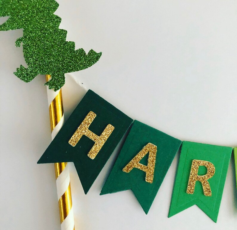 May include: Green and gold glitter banner with the word "Happy" spelled out. The banner is attached to a gold and white striped straw with a green glitter dinosaur on top.