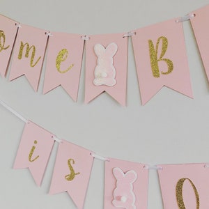 Op de afbeelding: Roze en gouden banner met de woorden "Home is" en een konijnenafbeelding. De banner is aan een wit lint geregen.
