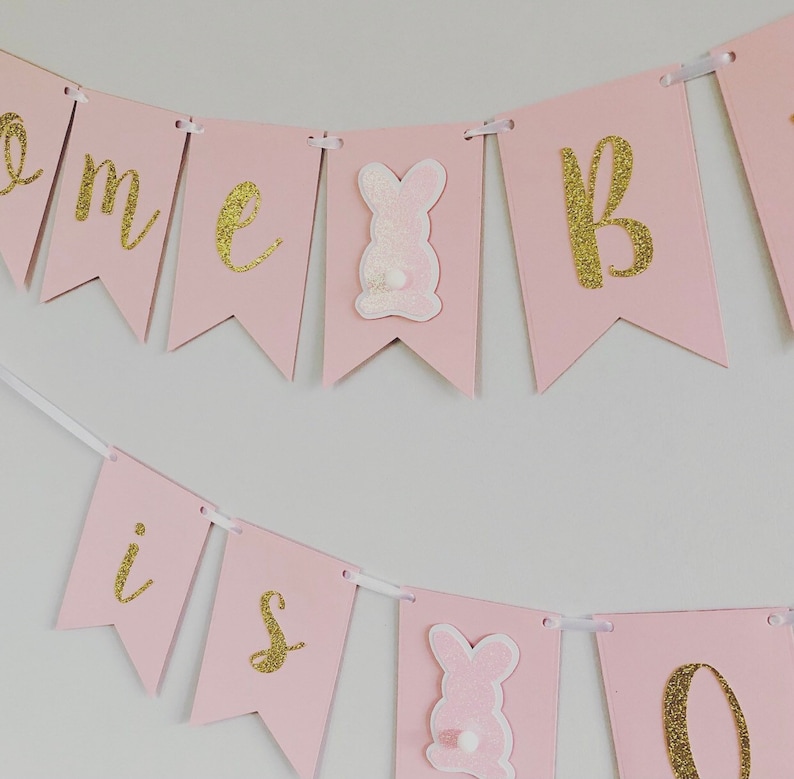 Op de afbeelding: Roze en gouden banner met de woorden "Home is" en een konijnenafbeelding. De banner is aan een wit lint geregen.