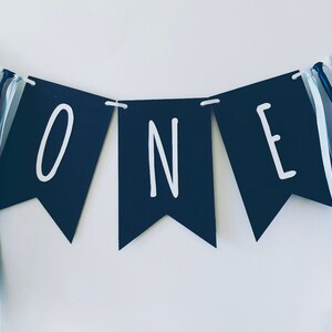 Puede incluir: Una pancarta negra y blanca con la palabra "ONE" en letras blancas. La pancarta est&aacute; decorada con cintas azules y blancas.
