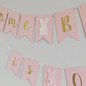 Op de afbeelding: Roze en gouden banner met de woorden "Come is one" en konijnen uitsnijdingen.