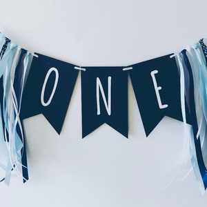 Puede incluir: Una pancarta azul marino con letras blancas que deletrean "ONE" con cintas azules y blancas colgando de cada extremo.