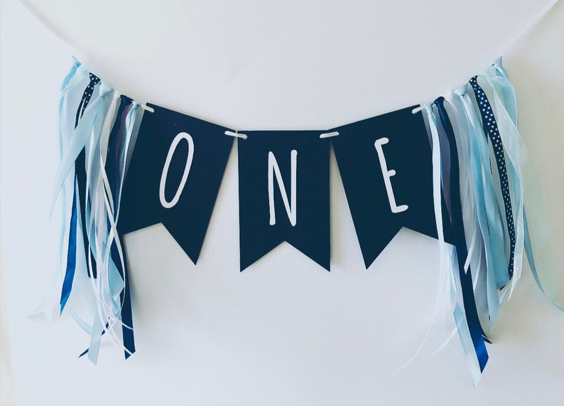 Puede incluir: Una pancarta azul marino con letras blancas que deletrean "ONE" con cintas azules y blancas colgando de cada extremo.