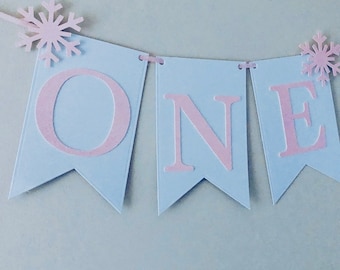 Puede incluir: Una pancarta azul claro con la palabra "ONE" en letras rosas. La pancarta está decorada con copos de nieve blancos.